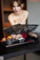 A naked woman sitting at a table holding a black fan.