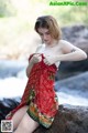 A woman in a red dress sitting on a rock by a river.