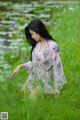 A woman in a pink dress sitting in the grass by a pond.