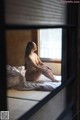 A naked woman sitting on a bed in front of a window.